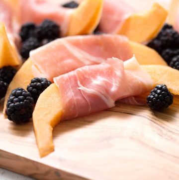 Cantaloupe slices wrapped in prosciutto with blackberries on a cutting board