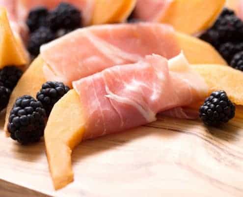 Cantaloupe slices wrapped in prosciutto with blackberries on a cutting board