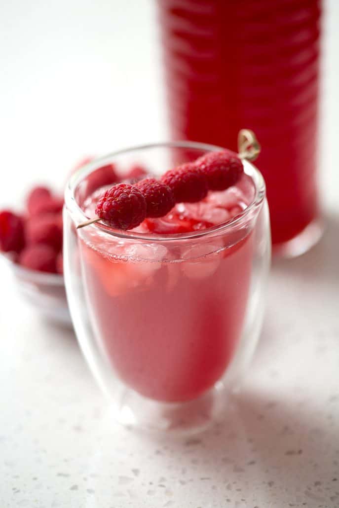 glass of raspberry thyme kombucha with toothpick of raspberries as a garnish on white background